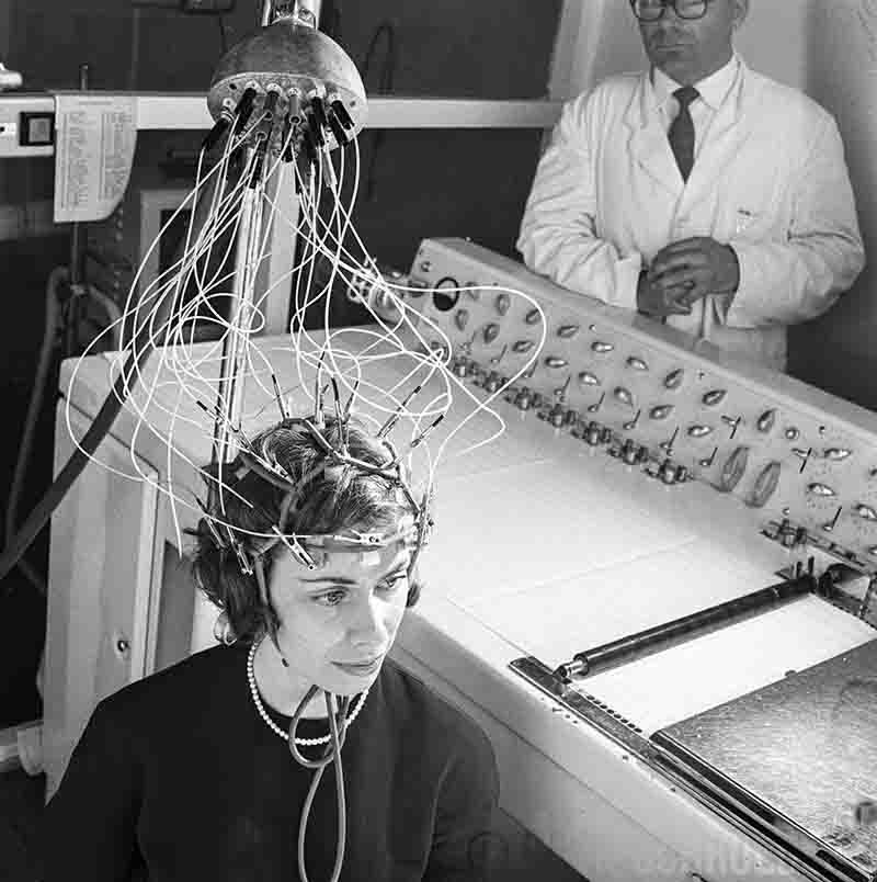 Black-and-white vintage photo of a woman wearing an EEG brainwave headset connected to a complex tangle of wires, with a scientist in a lab coat monitoring equipment in the background.