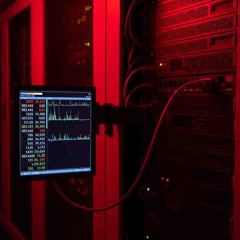 An AI server room is bathed in a dark red light. On the left side of the image, a monitor displays a graphical interface with lines and charts, showing real-time data and numbers in a list, all in white, green, and red text on a black background.