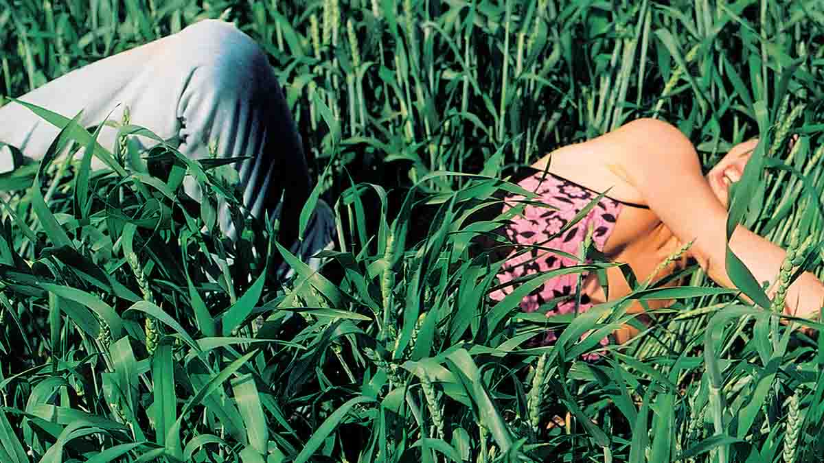 A person is lying down and partially hidden amidst a dense, vibrant green field of tall grass or young wheat. On the left, their bent knees are visible, covered in light gray or light blue trousers, symbolizing the need to step away from screens and reconnect with reality.